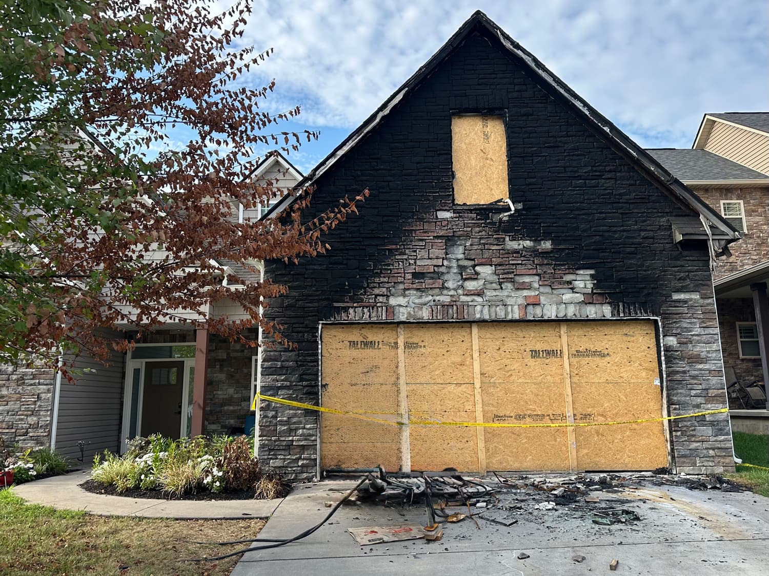 Exterior of a house with a fire-damaged garage showing blackened bricks, boarded-up doors, and caution tape after a residential fire.