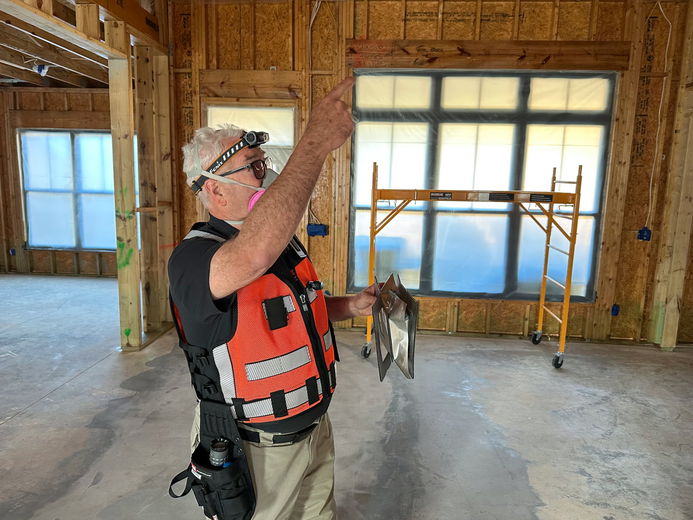 Inspector wearing a safety vest and respirator points toward the ceiling inside a framed building with covered windows and scaffolding